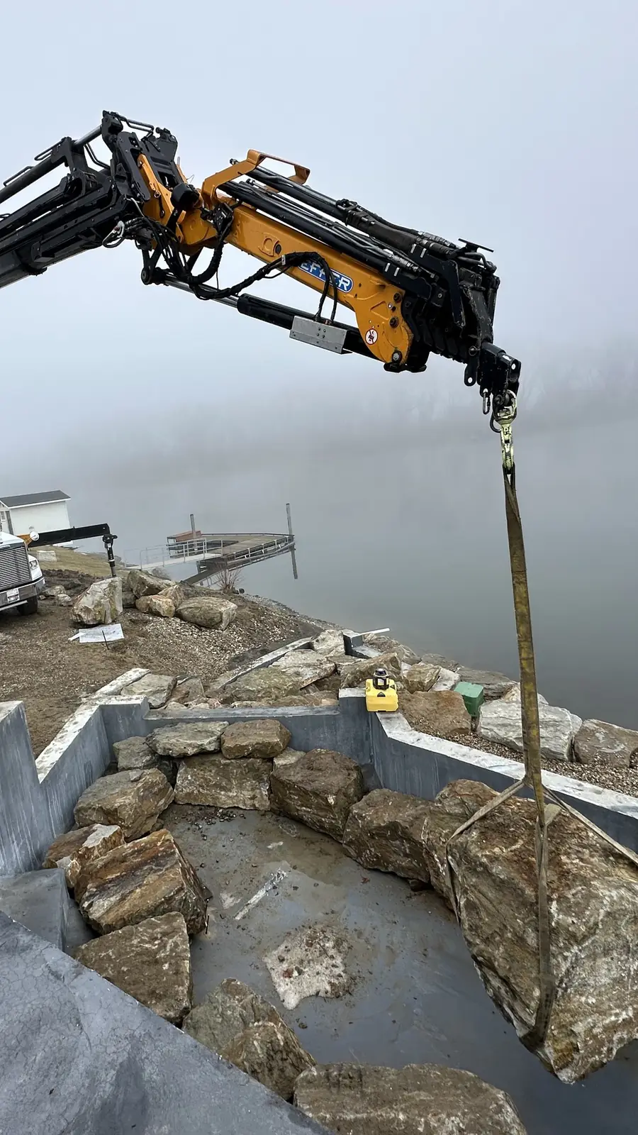 Parker knuckle-boom crane lowering a boulder onto a shoreline rock wall on a foggy day